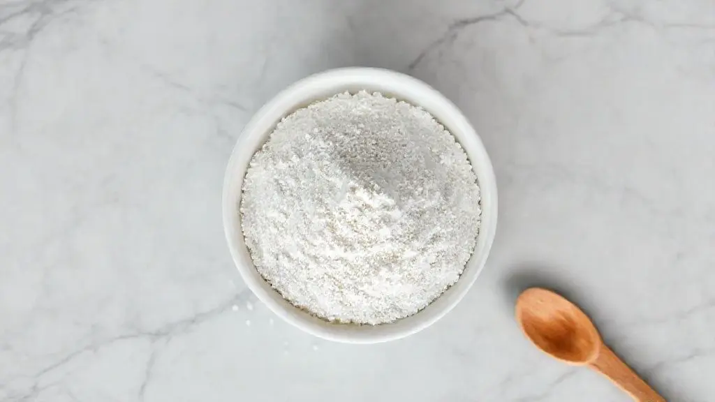 Suplemento de creatina em pó em uma tigela de cerâmica branca sobre superfície de mármore cinza, com colher de madeira ao lado.